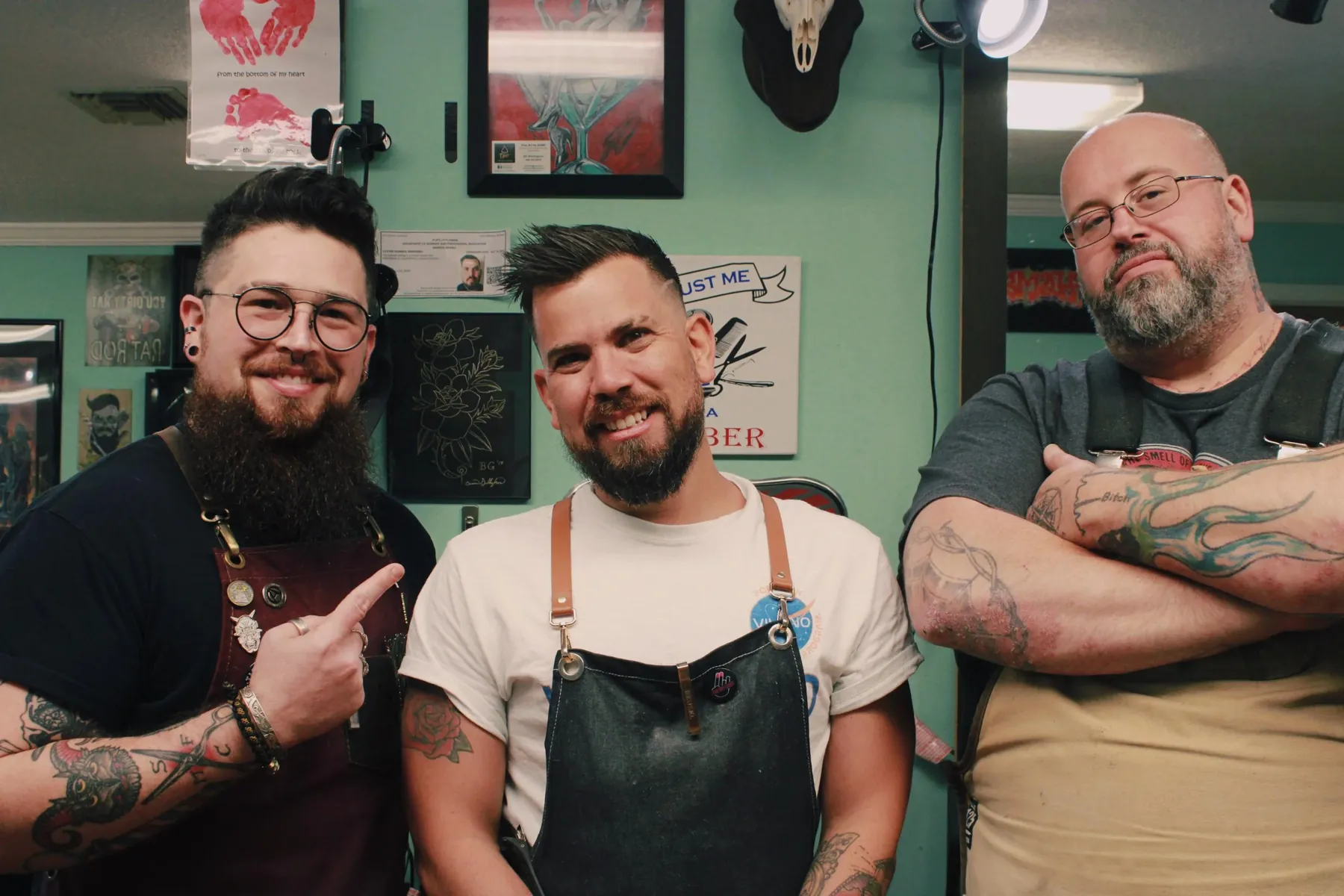 Three Fat Cats barbers standing together inside the shop, in front of the green wall of framed posters.